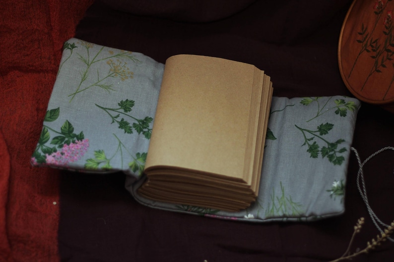 May include: An open journal with blank pages rests on a floral patterned fabric. The fabric is light gray with green and pink floral designs. The journal's pages are a light brown color.