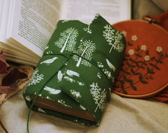 May include: A green fabric-covered journal with a white bird and tree pattern, tied with twine, and resting on a beige surface.