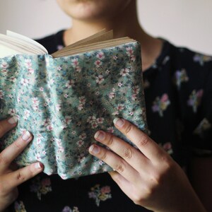 May include: A book with a floral fabric cover in shades of green, pink, and white. The book is open, revealing the pages. The cover is held by a person. The book cover has a floral pattern.
