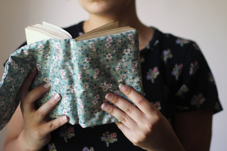May include: A book with a floral fabric cover in shades of green, pink, and white. The book is open, revealing the pages. The cover is held by a person. The book cover has a floral pattern.