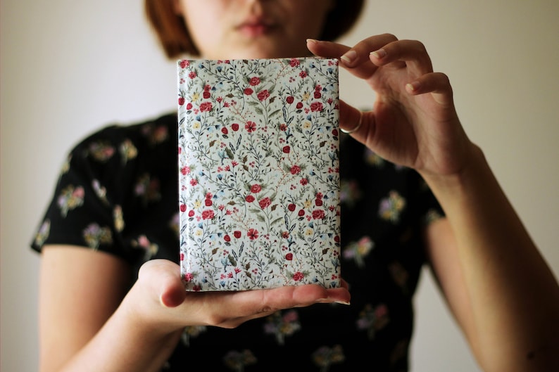 May include: A rectangular book cover with a floral pattern. The cover features a white background with a repeating pattern of red, yellow, and blue flowers, along with green leaves and stems. The book is held by a person.