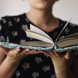 May include: An open book with aged pages, held in a floral fabric cover. The cover is light blue with a delicate floral pattern. The book's pages are yellowed with age, suggesting a vintage or antique item.