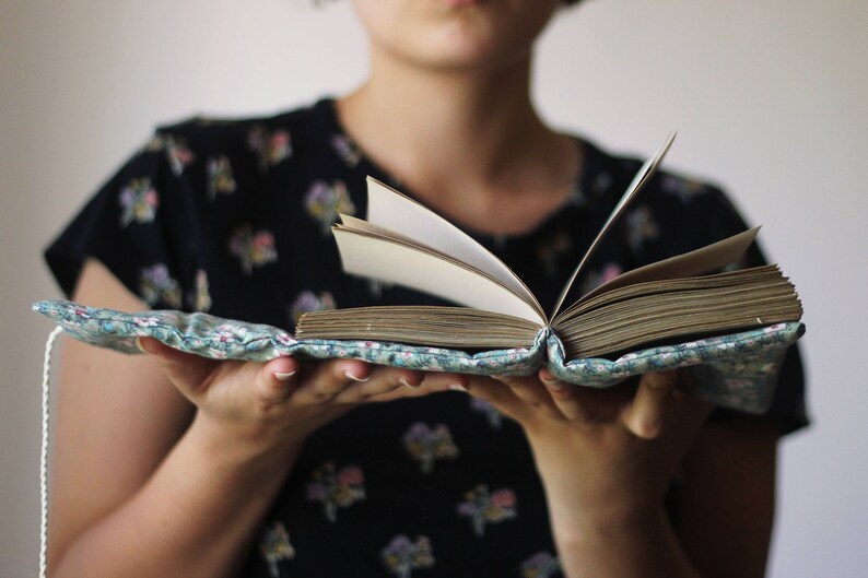 May include: An open book with aged pages, held in a floral fabric cover. The cover is light blue with a delicate floral pattern. The book's pages are yellowed with age, suggesting a vintage or antique item.