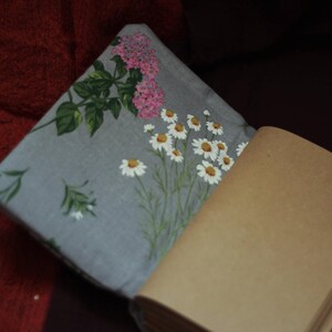 May include: A gray floral pillow with a daisy and pink flower design, next to a brown paper notebook. The pillow has a floral pattern with white daisies and pink flowers. The notebook has a stack of brown pages.