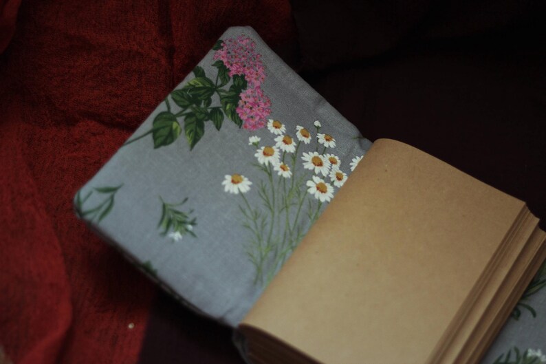 May include: A gray floral pillow with a daisy and pink flower design, next to a brown paper notebook. The pillow has a floral pattern with white daisies and pink flowers. The notebook has a stack of brown pages.