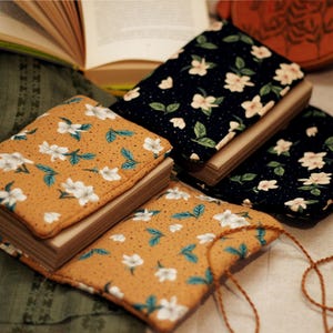May include: Books with floral covers in mustard yellow and navy blue. The covers feature white flowers and green or blue leaves. The books are open and closed, with a natural cord visible on one.