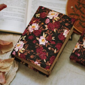 May include: Two black fabric-covered journals with a floral pattern in shades of pink, red, and brown. The journals are closed and lying on a white surface.