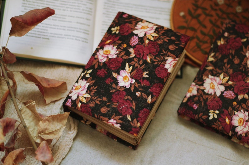May include: Two black fabric-covered journals with a floral pattern in shades of pink, red, and brown. The journals are closed and lying on a white surface.