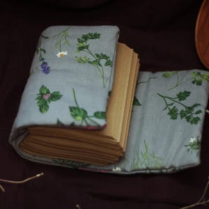 May include: A handmade journal with a light blue floral fabric cover featuring green leaves and white and purple flowers. The journal is open, revealing the pages inside. A white cord is attached to the cover.