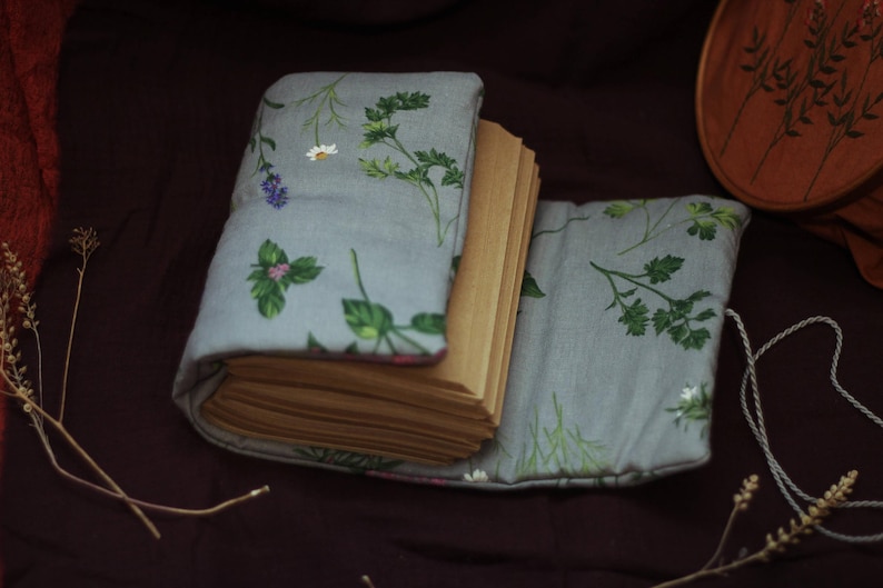 May include: A handmade journal with a light blue floral fabric cover featuring green leaves and white and purple flowers. The journal is open, revealing the pages inside. A white cord is attached to the cover.