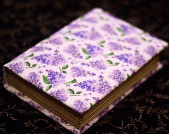 Puede incluir: Un libro de tapa dura con un diseño floral de lilas en la portada. La portada presenta un patrón de flores de lilas moradas y hojas verdes sobre un fondo blanco. Las páginas del libro son de color marrón dorado.