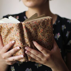 May include: An open book with pages visible, held by a person. The book has a floral patterned cover in shades of tan, red, and orange. The person is wearing a black floral print top.