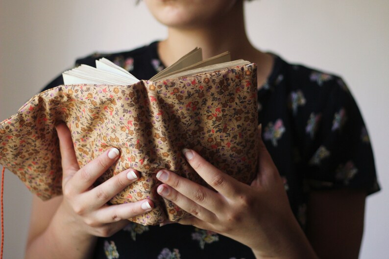 May include: An open book with pages visible, held by a person. The book has a floral patterned cover in shades of tan, red, and orange. The person is wearing a black floral print top.