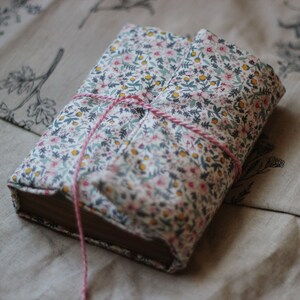 May include: A hardcover book wrapped in a floral fabric cover with pink string tied around it. The fabric features a pattern of small pink, yellow, and blue flowers on a white background. The book is resting on a beige fabric with botanical illustrations.