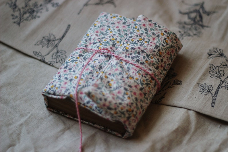 May include: A hardcover book wrapped in a floral fabric cover with pink string tied around it. The fabric features a pattern of small pink, yellow, and blue flowers on a white background. The book is resting on a beige fabric with botanical illustrations.