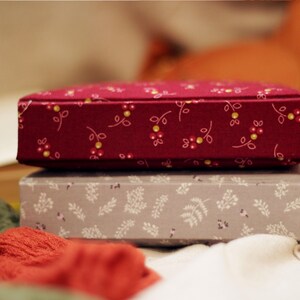 May include: Two decorative boxes stacked on top of each other. The top box is a deep red with a floral pattern of small white and yellow flowers. The bottom box is gray with a white leaf pattern. The boxes are on a white surface.