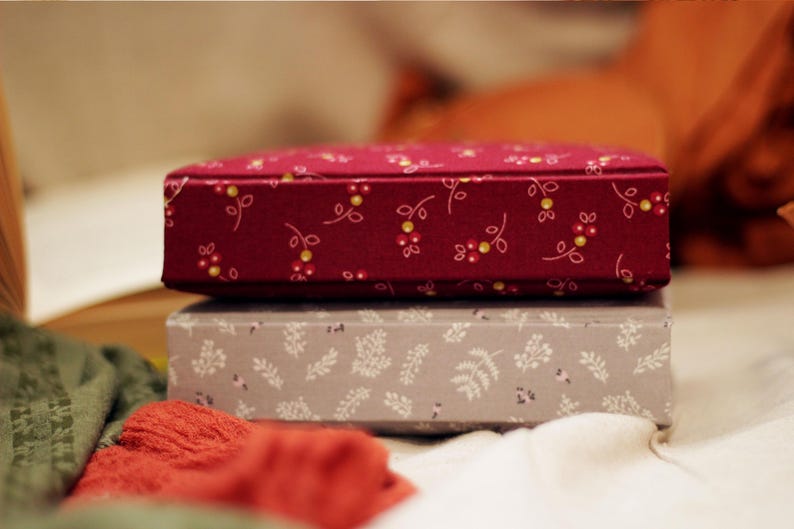 May include: Two decorative boxes stacked on top of each other. The top box is a deep red with a floral pattern of small white and yellow flowers. The bottom box is gray with a white leaf pattern. The boxes are on a white surface.