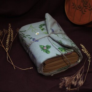 May include: A bound book wrapped in a light blue fabric cover with a floral pattern of green leaves and purple flowers. The book is tied with twine. Dried plants are placed around the book on a dark brown surface.