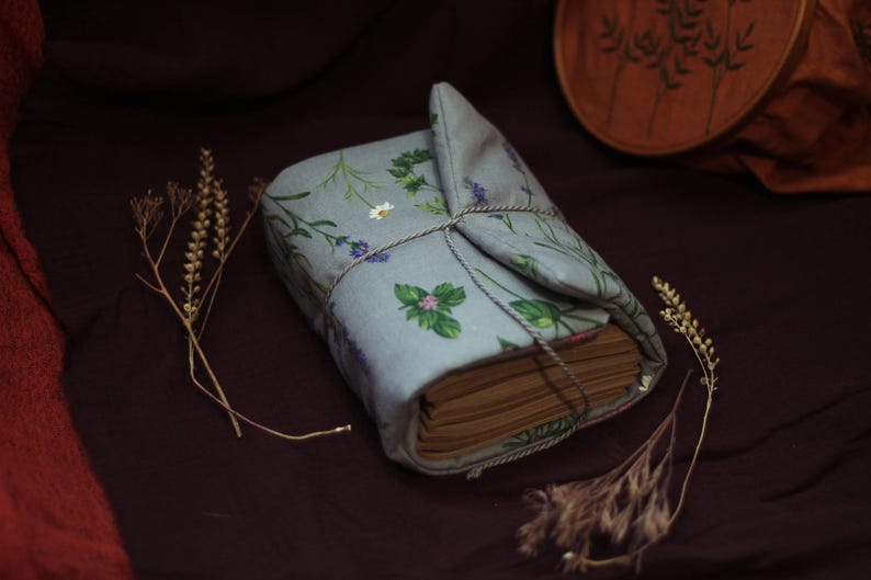 May include: A bound book wrapped in a light blue fabric cover with a floral pattern of green leaves and purple flowers. The book is tied with twine. Dried plants are placed around the book on a dark brown surface.