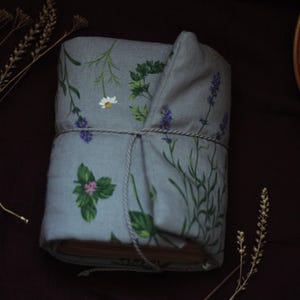 May include: A wrapped book with a light blue floral fabric cover featuring illustrations of herbs and flowers, including lavender, daisies, and basil. The book is tied with a natural twine, and sprigs of dried flowers are placed around it.