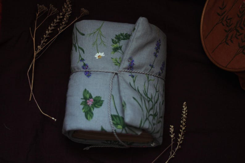 May include: A wrapped book with a light blue floral fabric cover featuring illustrations of herbs and flowers, including lavender, daisies, and basil. The book is tied with a natural twine, and sprigs of dried flowers are placed around it.