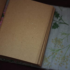 May include: An open journal with blank, brown pages. The journal is lying on a light blue fabric with a floral pattern in green and beige. The journal's spine is visible, with a floral design.