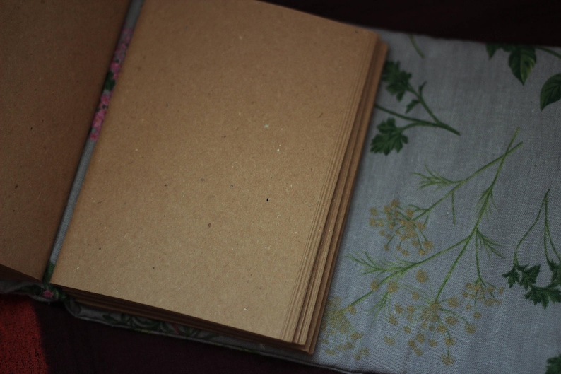 May include: An open journal with blank, brown pages. The journal is lying on a light blue fabric with a floral pattern in green and beige. The journal's spine is visible, with a floral design.