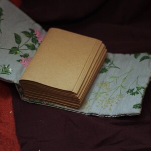 May include: A handmade journal with a stack of blank pages. The journal is encased in a light blue fabric cover adorned with floral patterns in green and pink. A white string is attached.