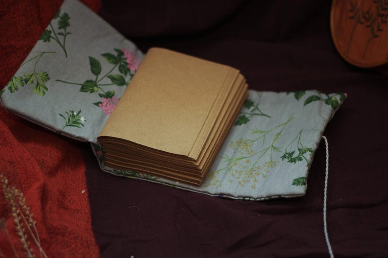 May include: A handmade journal with a stack of blank pages. The journal is encased in a light blue fabric cover adorned with floral patterns in green and pink. A white string is attached.