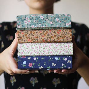 Puede incluir: Una pila de cuatro cajas rectangulares con cubiertas de tela floral. Las cajas se sostienen frente a una persona. Los patrones de tela incluyen verde azulado, marr&oacute;n, blanco y azul marino con varios dise&ntilde;os de flores y mariposas.