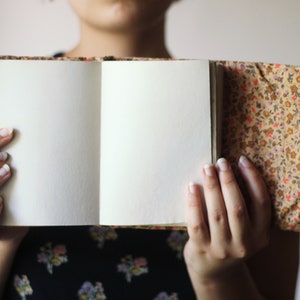May include: An open journal with blank, cream-colored pages. The cover and spine are adorned with a floral pattern in shades of brown, orange, and pink. An orange string is attached to the journal. The journal is held open.