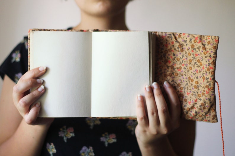 May include: An open journal with blank, cream-colored pages. The cover and spine are adorned with a floral pattern in shades of brown, orange, and pink. An orange string is attached to the journal. The journal is held open.
