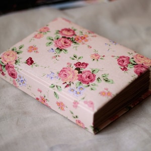 Puede incluir: Una caja rectangular cerrada con un estampado floral de rosas rosas, detalles rojos y flores azules sobre un fondo rosa claro. La caja tiene una est&eacute;tica vintage y est&aacute; sobre una superficie de color neutro.