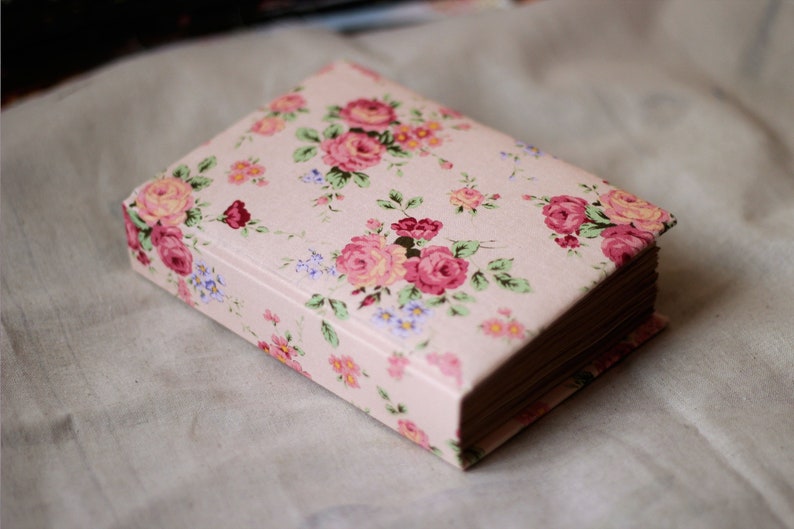 Puede incluir: Una caja rectangular cerrada con un estampado floral de rosas rosas, detalles rojos y flores azules sobre un fondo rosa claro. La caja tiene una est&eacute;tica vintage y est&aacute; sobre una superficie de color neutro.