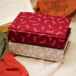 May include: Two fabric-covered boxes stacked on top of each other. The top box is a deep red with a floral pattern in yellow and white. The bottom box is gray with a white leaf pattern. An open book and fabric are in the background.