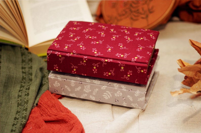 May include: Two fabric-covered boxes stacked on top of each other. The top box is a deep red with a floral pattern in yellow and white. The bottom box is gray with a white leaf pattern. An open book and fabric are in the background.