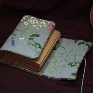 May include: A small, open book with aged pages, nestled in a light blue floral fabric cover. The cover features a pattern of white daisies, pink flowers, and green foliage. The book is on a dark maroon surface.