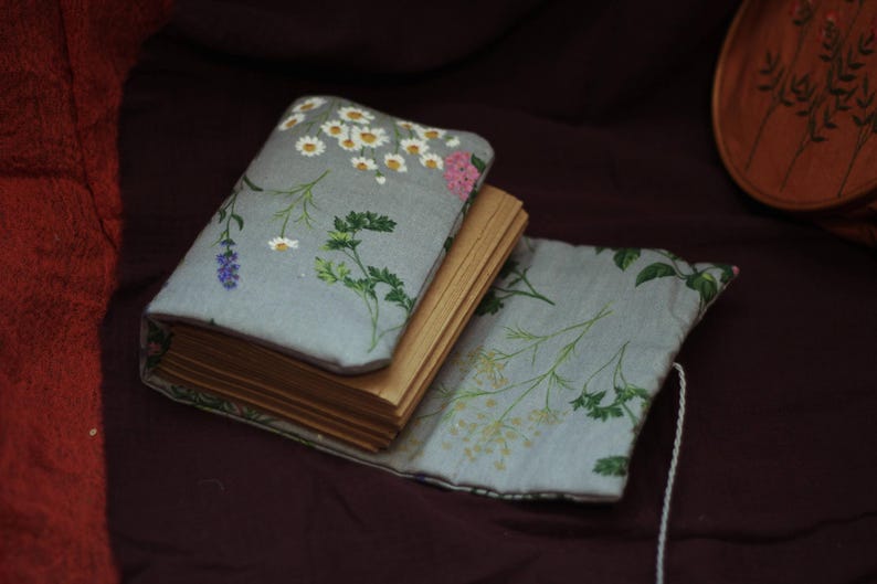 May include: A small, open book with aged pages, nestled in a light blue floral fabric cover. The cover features a pattern of white daisies, pink flowers, and green foliage. The book is on a dark maroon surface.
