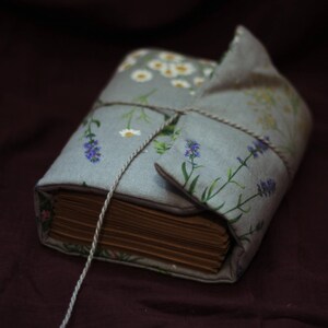 May include: A bound book wrapped in a light blue floral fabric cover, featuring lavender and daisy patterns. The book is tied with a thin, light-colored string. The pages are visible and have a warm, aged tone.