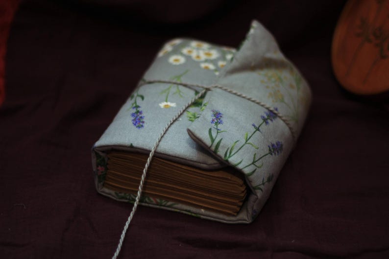 May include: A bound book wrapped in a light blue floral fabric cover, featuring lavender and daisy patterns. The book is tied with a thin, light-colored string. The pages are visible and have a warm, aged tone.