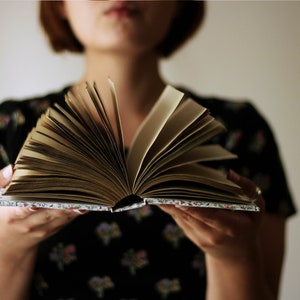 May include: An open book with aged, yellowed pages is held in front of a person. The book's cover has a floral pattern. The person is wearing a black dress with a floral pattern. The background is a plain, light color.