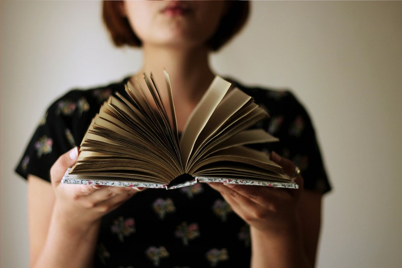 May include: An open book with aged, yellowed pages is held in front of a person. The book's cover has a floral pattern. The person is wearing a black dress with a floral pattern. The background is a plain, light color.
