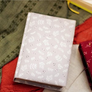 May include: A light gray journal with a white floral pattern, next to a burgundy journal with a yellow and white floral pattern. The journals are on a red and green patterned fabric.