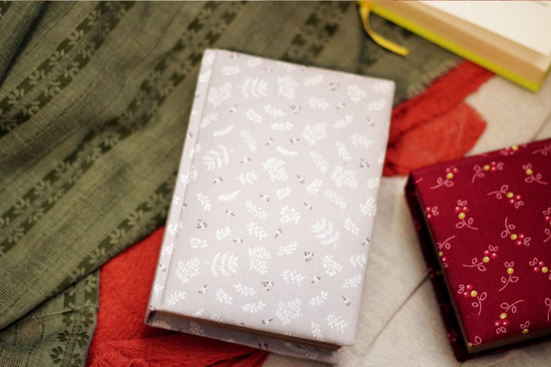 May include: A light gray journal with a white floral pattern, next to a burgundy journal with a yellow and white floral pattern. The journals are on a red and green patterned fabric.