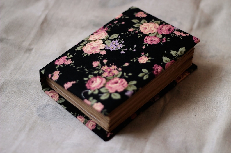 Puede incluir: Un libro de tapa dura con una cubierta de tela floral negra con rosas rosas y melocot&oacute;n, hojas verdes y acentos morados. Las p&aacute;ginas del libro son visibles y la est&eacute;tica general es vintage.
