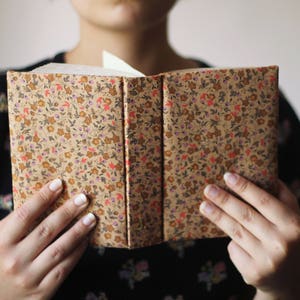 Puede incluir: Libro abierto con una cubierta estampada floral en tonos marr&oacute;n, rojo y amarillo. El libro se sostiene abierto, revelando las p&aacute;ginas blancas en el interior. La cubierta tiene un patr&oacute;n repetido de peque&ntilde;as flores y hojas.