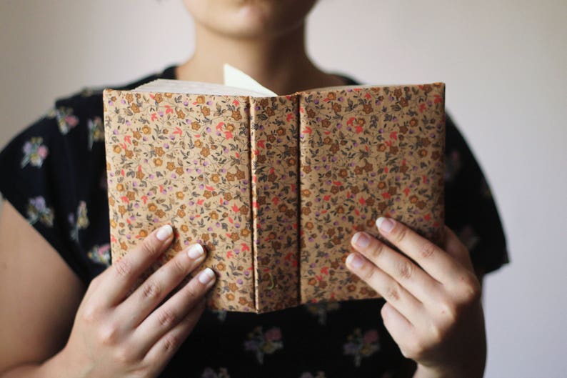 Puede incluir: Libro abierto con una cubierta estampada floral en tonos marr&oacute;n, rojo y amarillo. El libro se sostiene abierto, revelando las p&aacute;ginas blancas en el interior. La cubierta tiene un patr&oacute;n repetido de peque&ntilde;as flores y hojas.