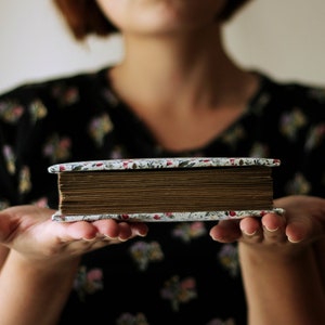 May include: A vintage-style photo album with a floral patterned cover and aged, brown-toned pages. The album is held in open hands, showcasing its rectangular shape and the detailed design of the cover. The album's spine is visible, suggesting a well-preserved keepsake.