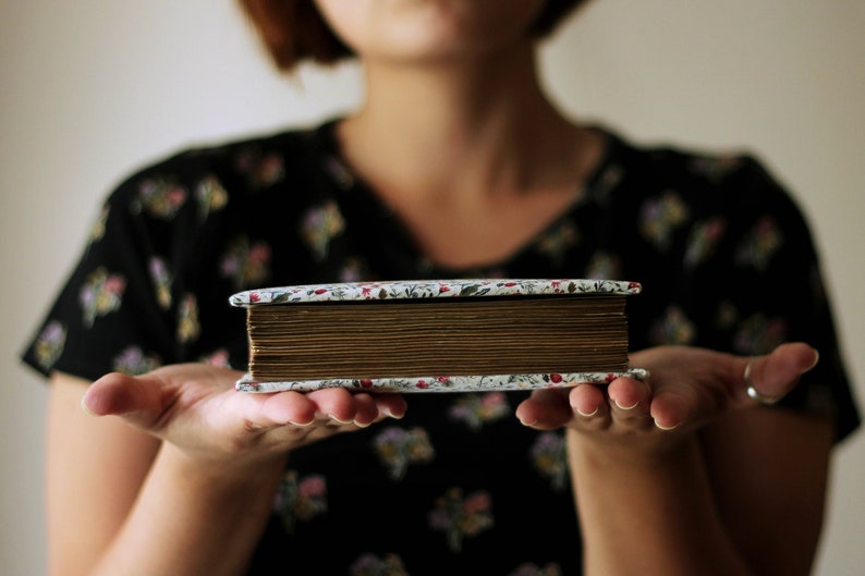 May include: A vintage-style photo album with a floral patterned cover and aged, brown-toned pages. The album is held in open hands, showcasing its rectangular shape and the detailed design of the cover. The album's spine is visible, suggesting a well-preserved keepsake.