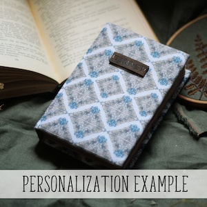 Puede incluir: Un libro cerrado con una cubierta estampada con un dise&ntilde;o geom&eacute;trico en tonos grises y azules. Una peque&ntilde;a etiqueta de madera con la palabra "TRAVEL" est&aacute; adjunta. El texto "PERSONALIZATION EXAMPLE" se muestra en la parte inferior de la imagen.